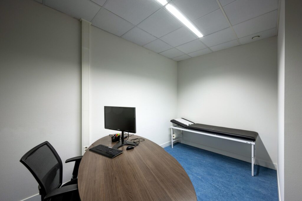 Een strak ingerichte dokterskamer in Laan van de Helende Meesters met bureau, computer en onderzoeksbank.